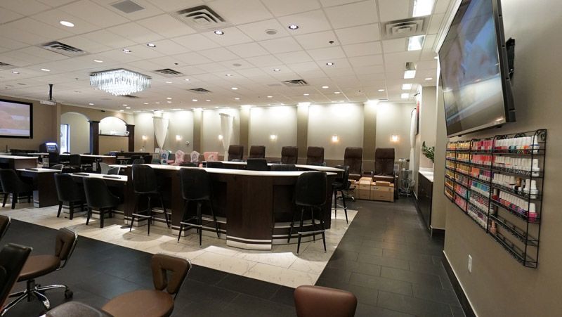 Interior of a nail salon with dark chairs, a central bar, and a wall-mounted TV.