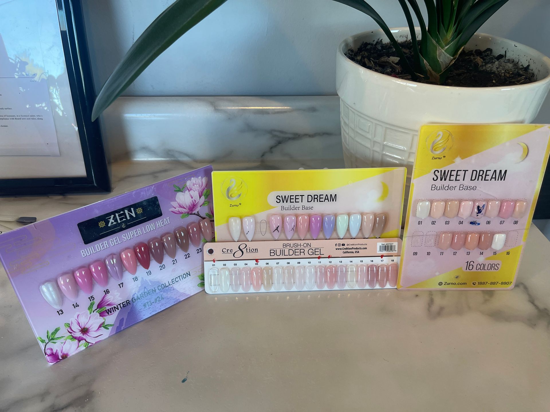 Various press-on nail kits in pastel colors on a marble surface, near a potted plant and framed artwork.