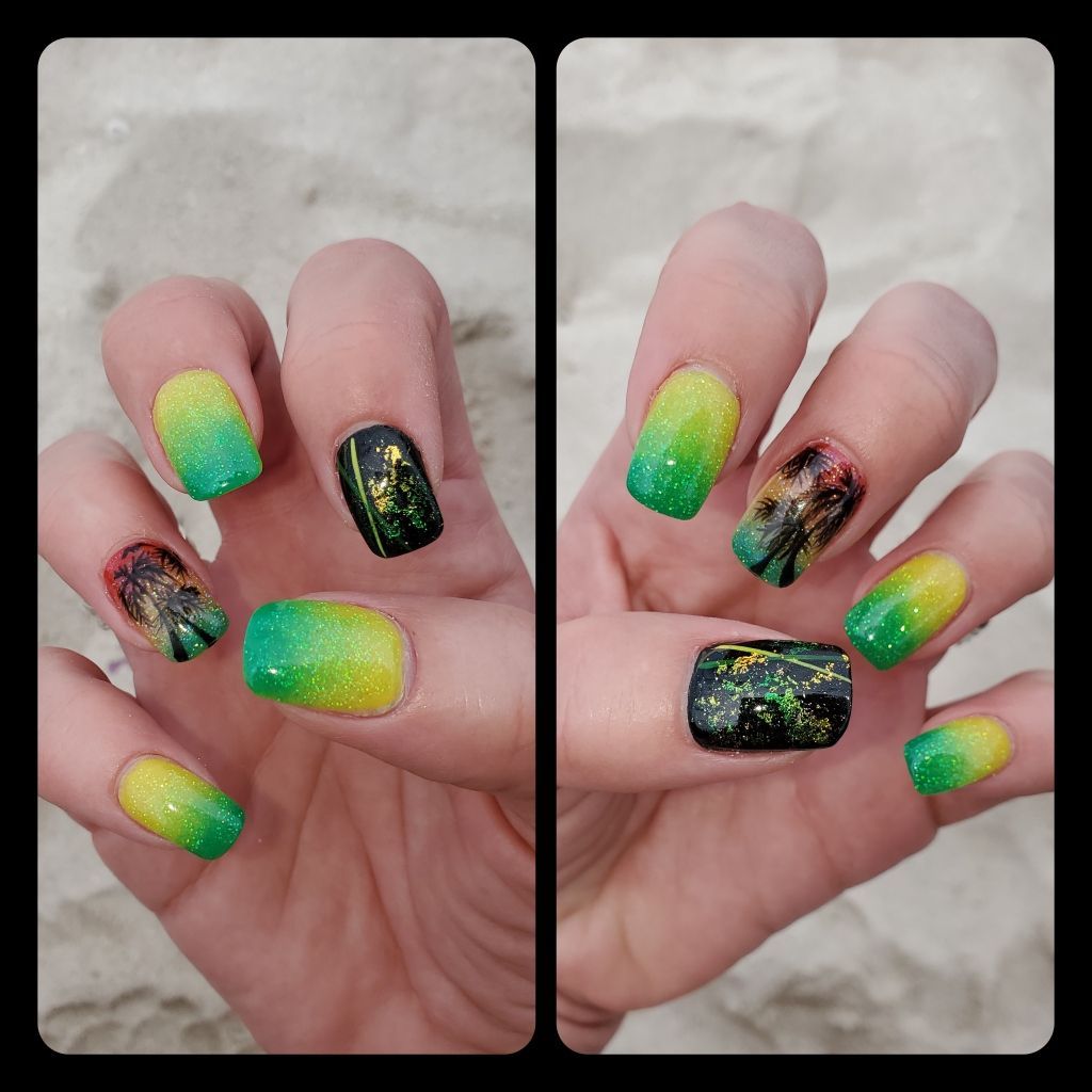 Hands with colorful nail art, including ombre yellow to green and black designs with gold accents, on a sandy surface.