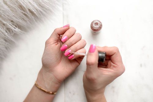 Hands applying clear glitter nail polish over bright pink nail polish.