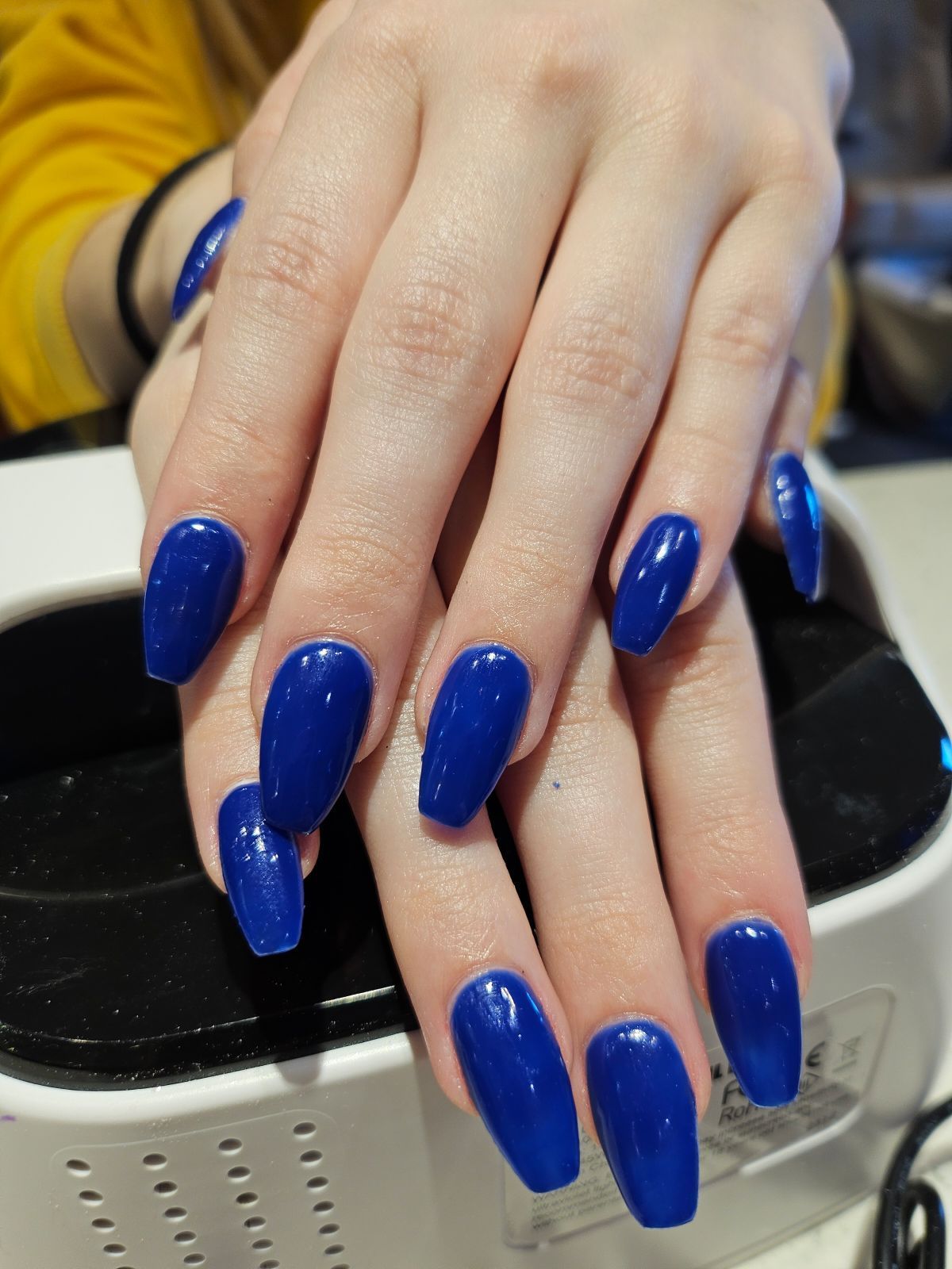 Hands with long, glossy blue coffin-shaped nails, resting on a nail dryer.