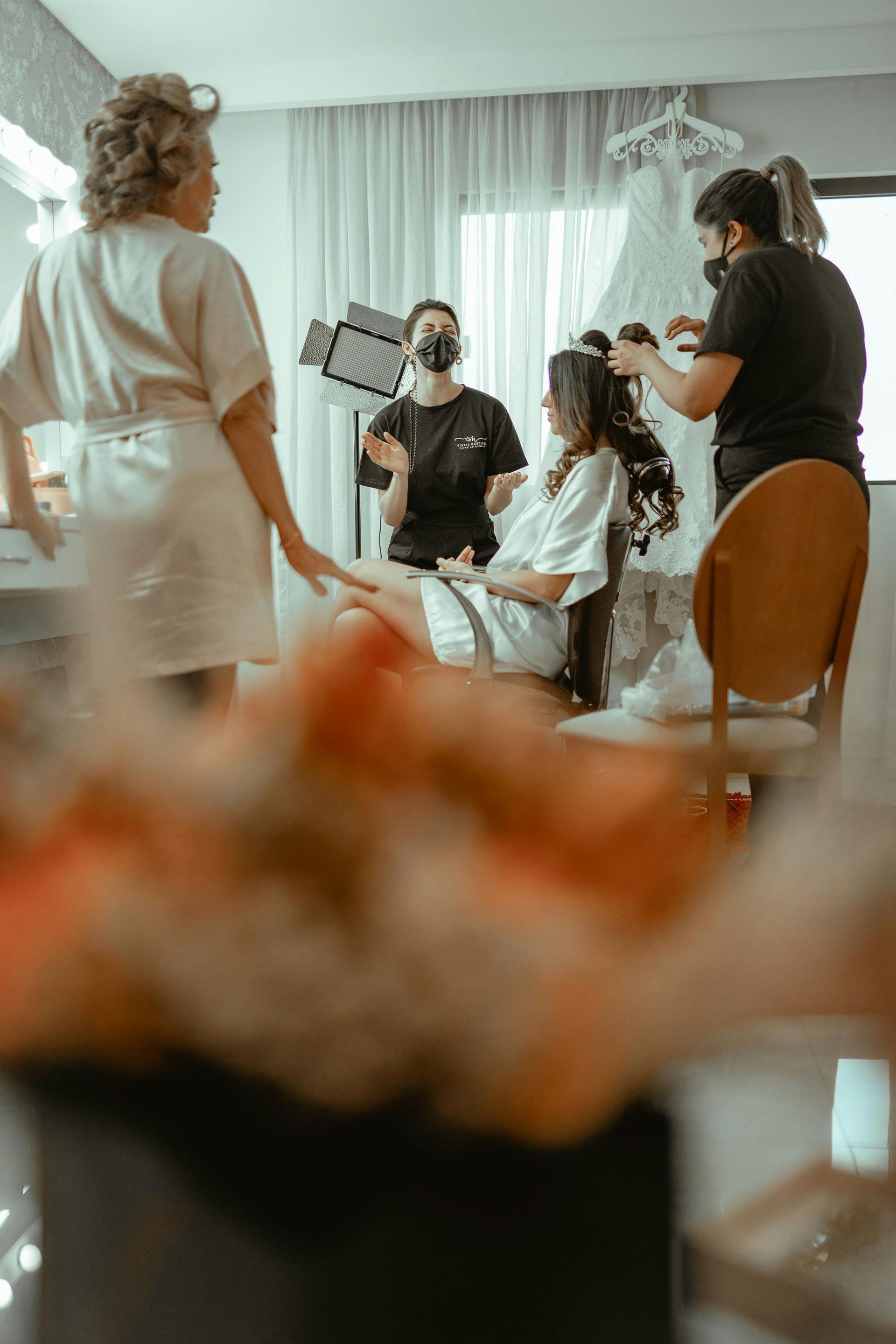 Bride getting her hair and makeup done, surrounded by a team, in a preparation room.