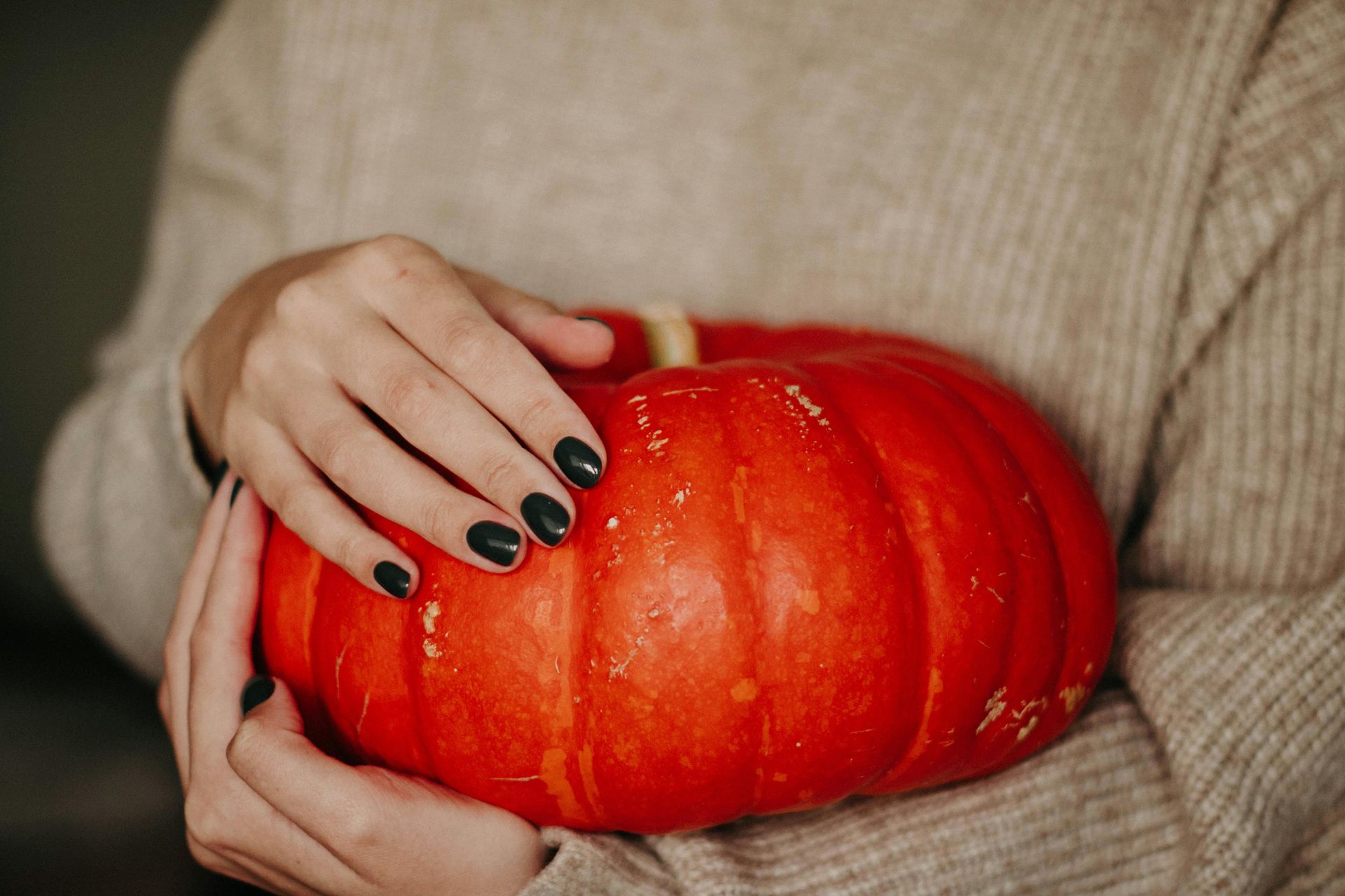 Person holding a small, bright orange pumpkin; they have black fingernails and are wearing a beige sweater.