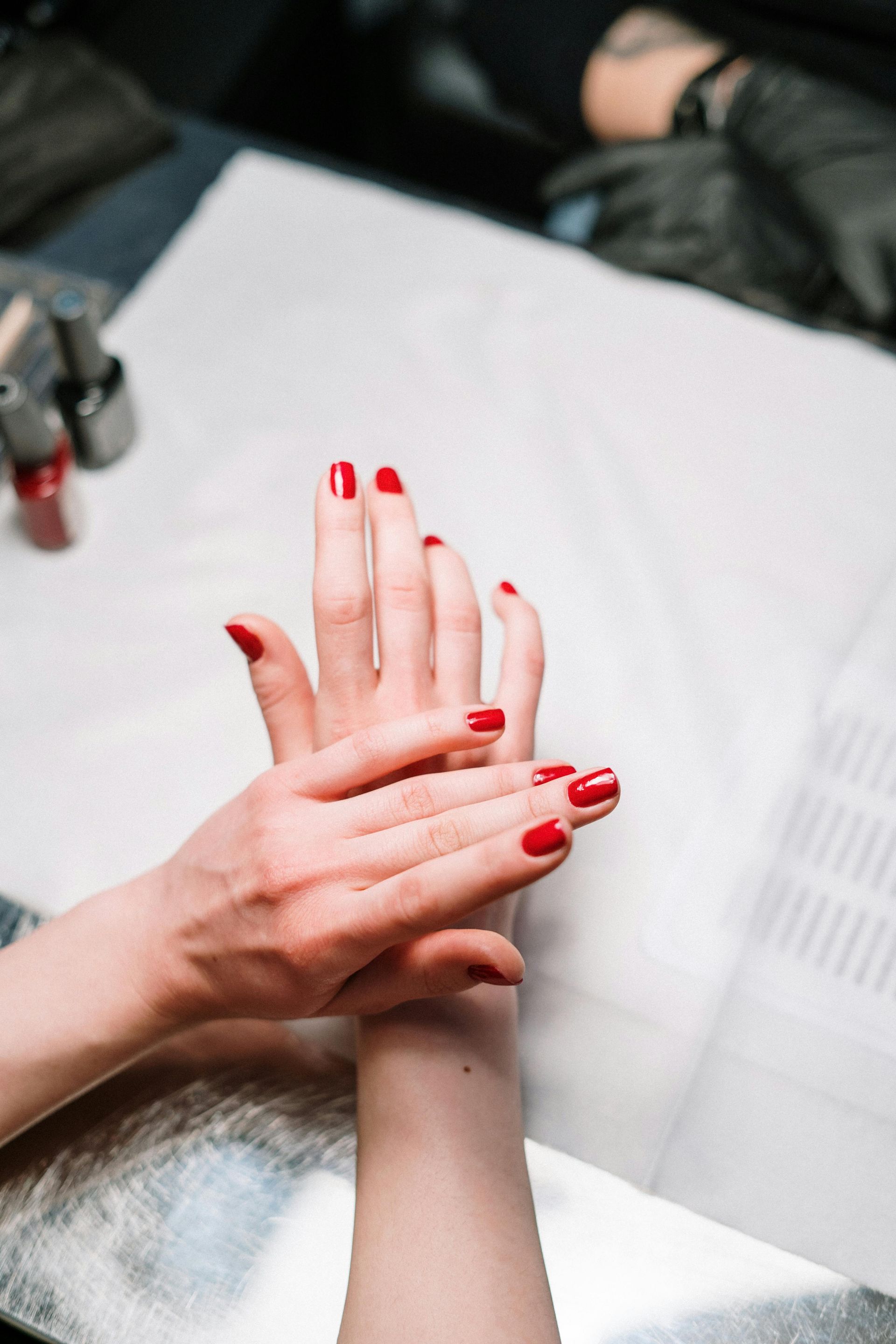 Hands with red painted nails being touched during a manicure on a white surface.