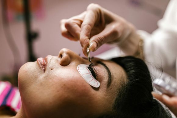 Woman getting eyelash extensions; technician using tweezers, eye patches, close-up.