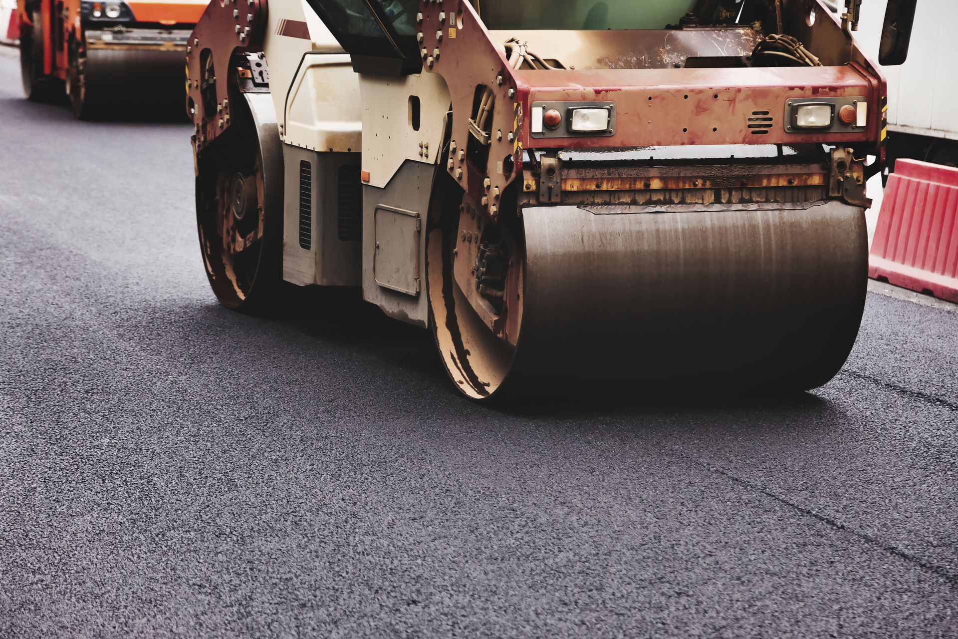 Road roller compacting fresh asphalt during street paving, with construction barriers nearby.
