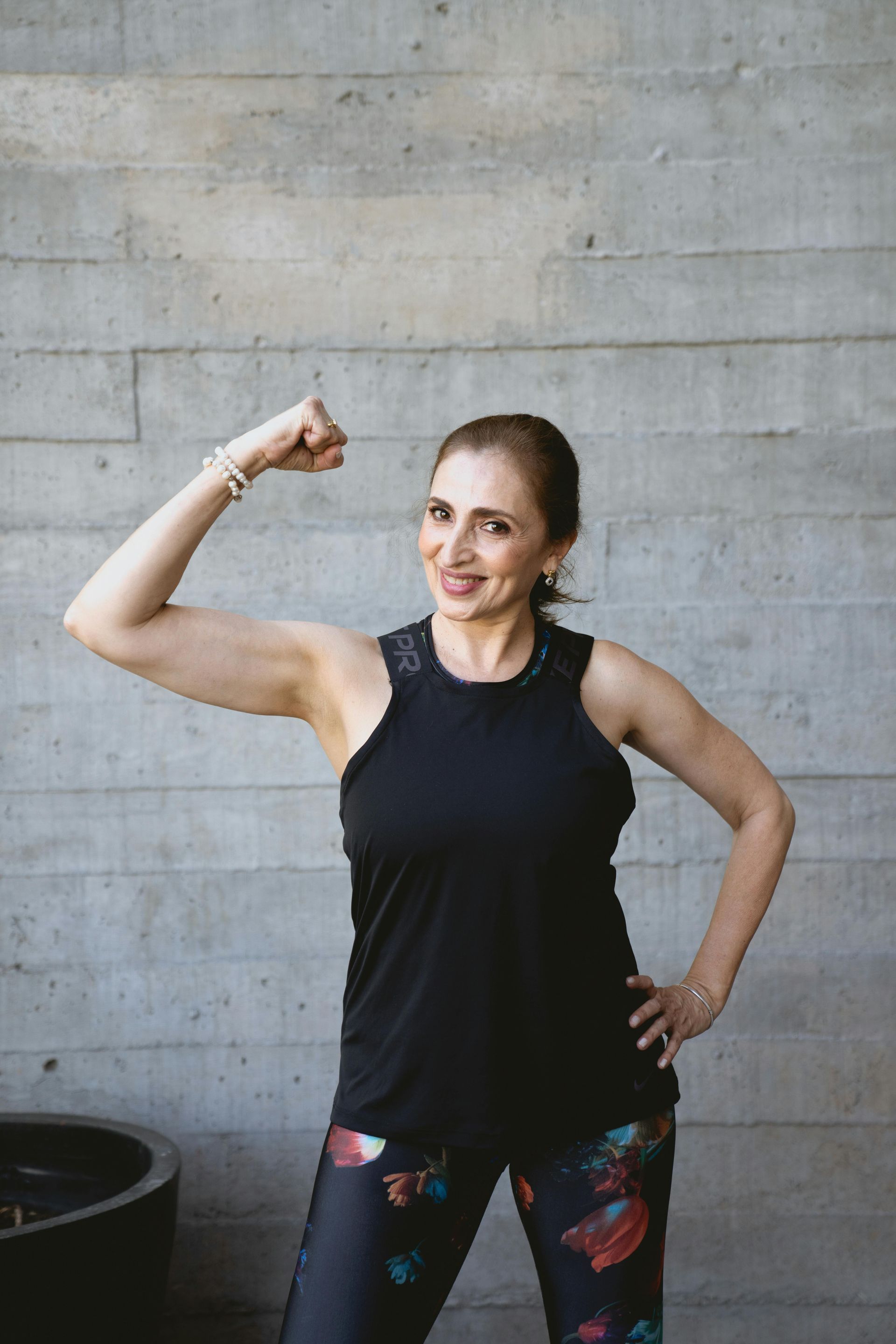 Woman flexing arm, smiling, wearing black tank top and patterned leggings against concrete wall.