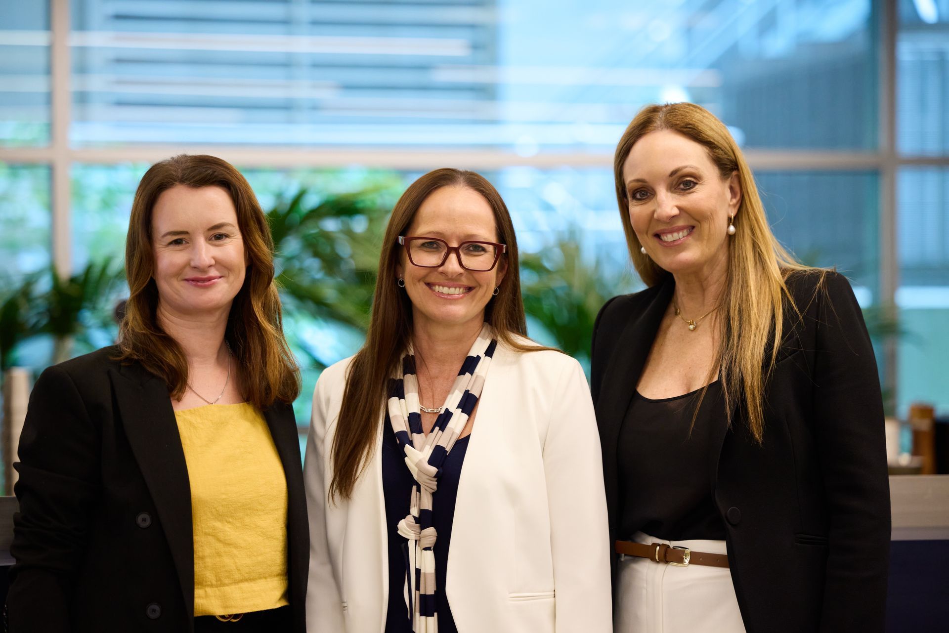 Three women in business attire pose in an office setting.
