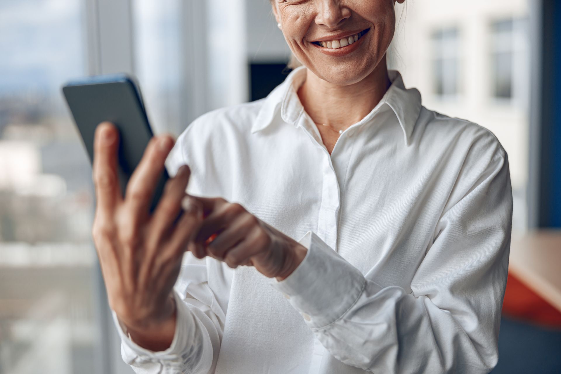 A woman in a white shirt is smiling while using a cell phone.