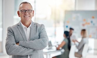 A man in a suit and glasses is standing with his arms crossed in an office.