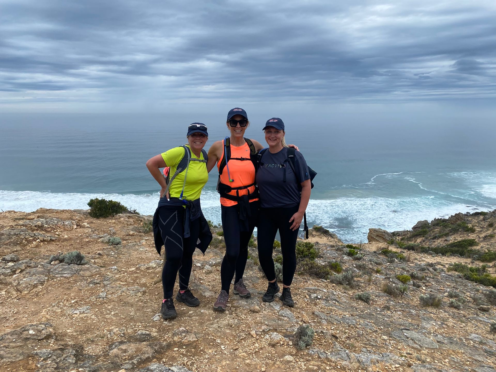 Three women are posing for a picture on top of a rocky hill near the ocean.