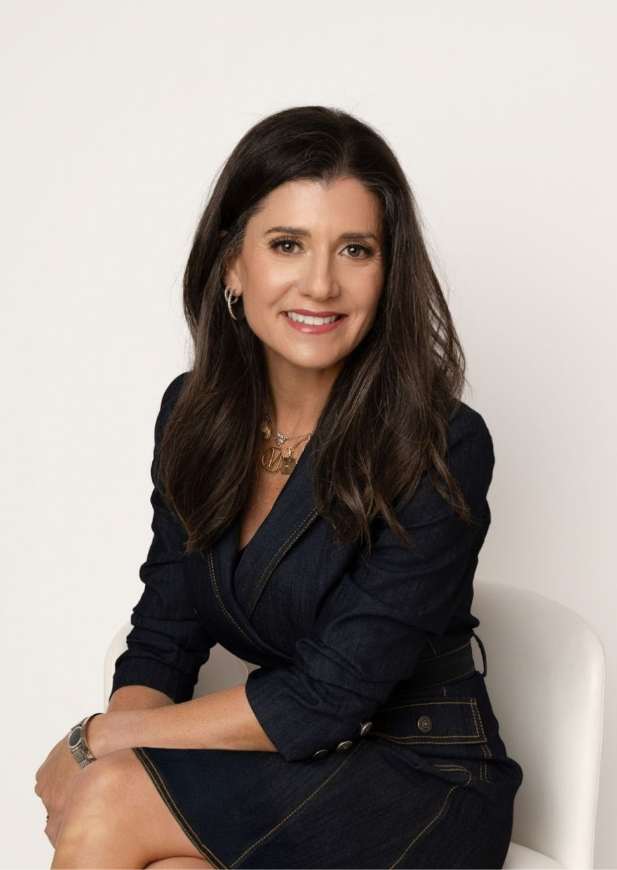 Woman in a dark denim jacket and skirt sits smiling, against a white backdrop.