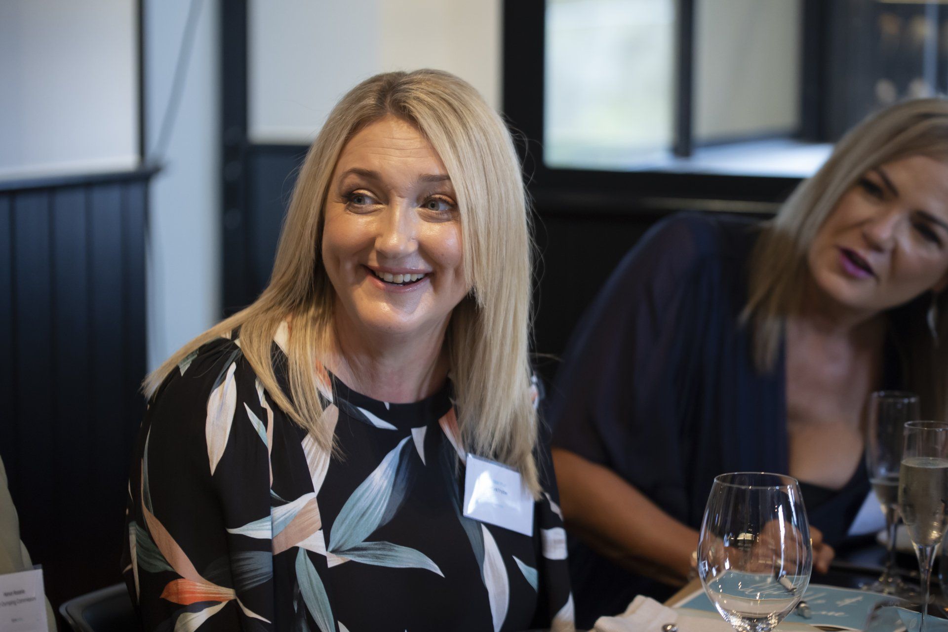 Two women are sitting at a table with wine glasses and smiling.