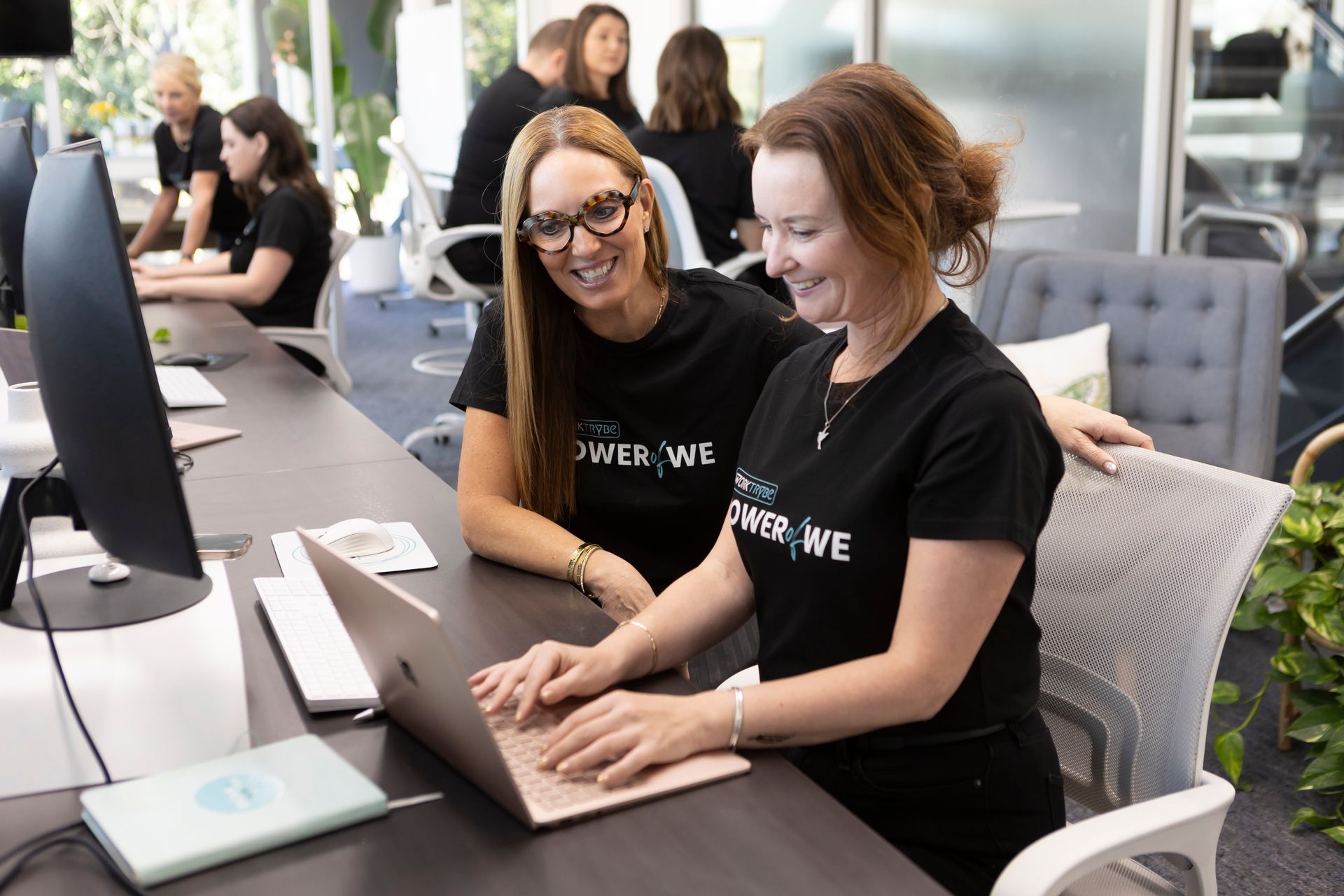 Two women are sitting at a desk looking at a laptop computer.