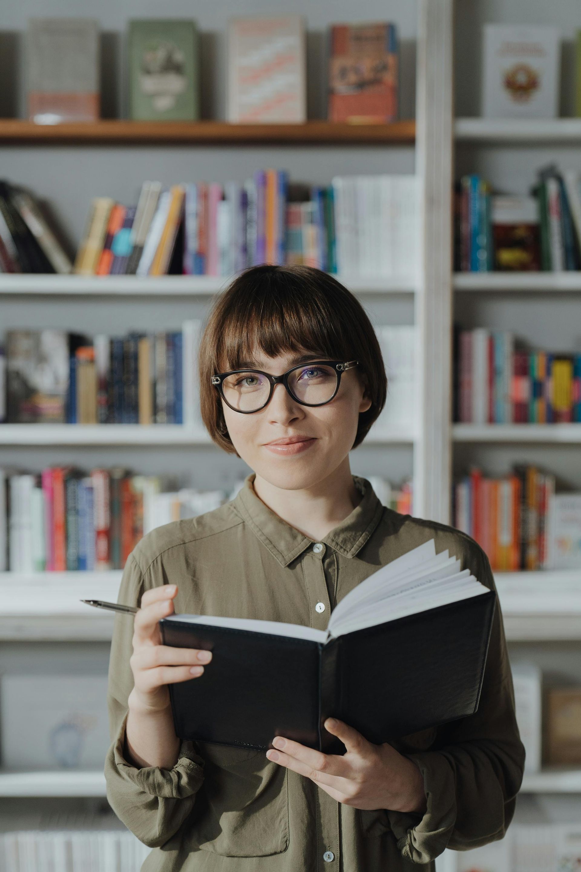 A woman wearing glasses is holding a book in a library.