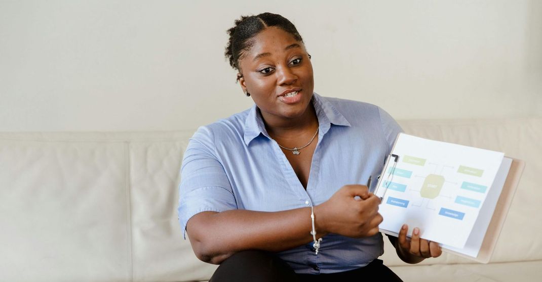 A woman is sitting on a couch holding a clipboard and a notebook.
