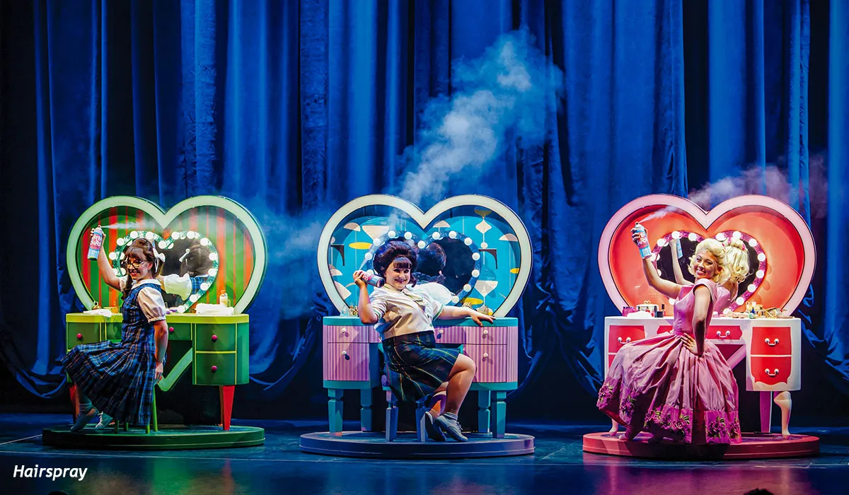 Three performers in front of heart-shaped vanities spraying hairspray on stage, lit with colorful spotlights.