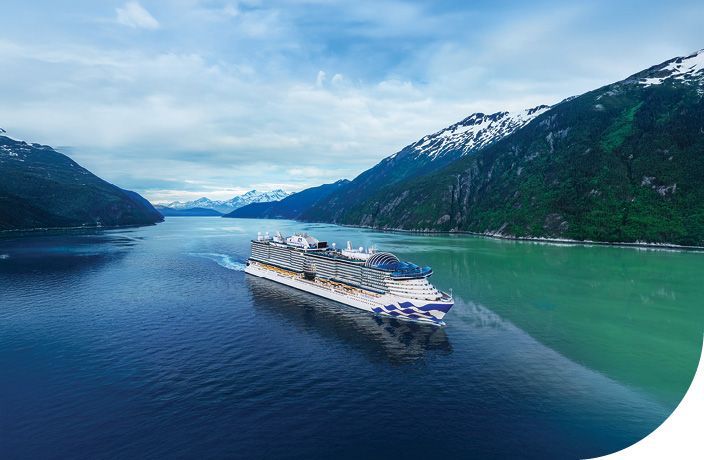 Cruise ship sailing through a blue fjord between green, snow-dusted mountains under a cloudy sky