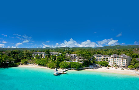 Beachfront resort with turquoise water, white sand, and buildings nestled in lush greenery under a blue sky.