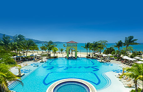 Large resort pool with ocean view, palm trees, and clear blue sky.