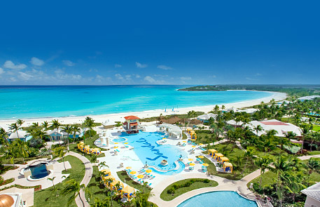 Aerial view of a tropical resort with turquoise ocean, white sand beach, and pools surrounded by lush green landscaping.
