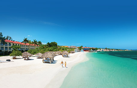 Beach resort with white sand, turquoise water, thatched huts, and couple walking.