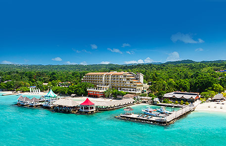 Oceanfront resort with buildings, docks, and clear turquoise water. Lush green trees cover the background.