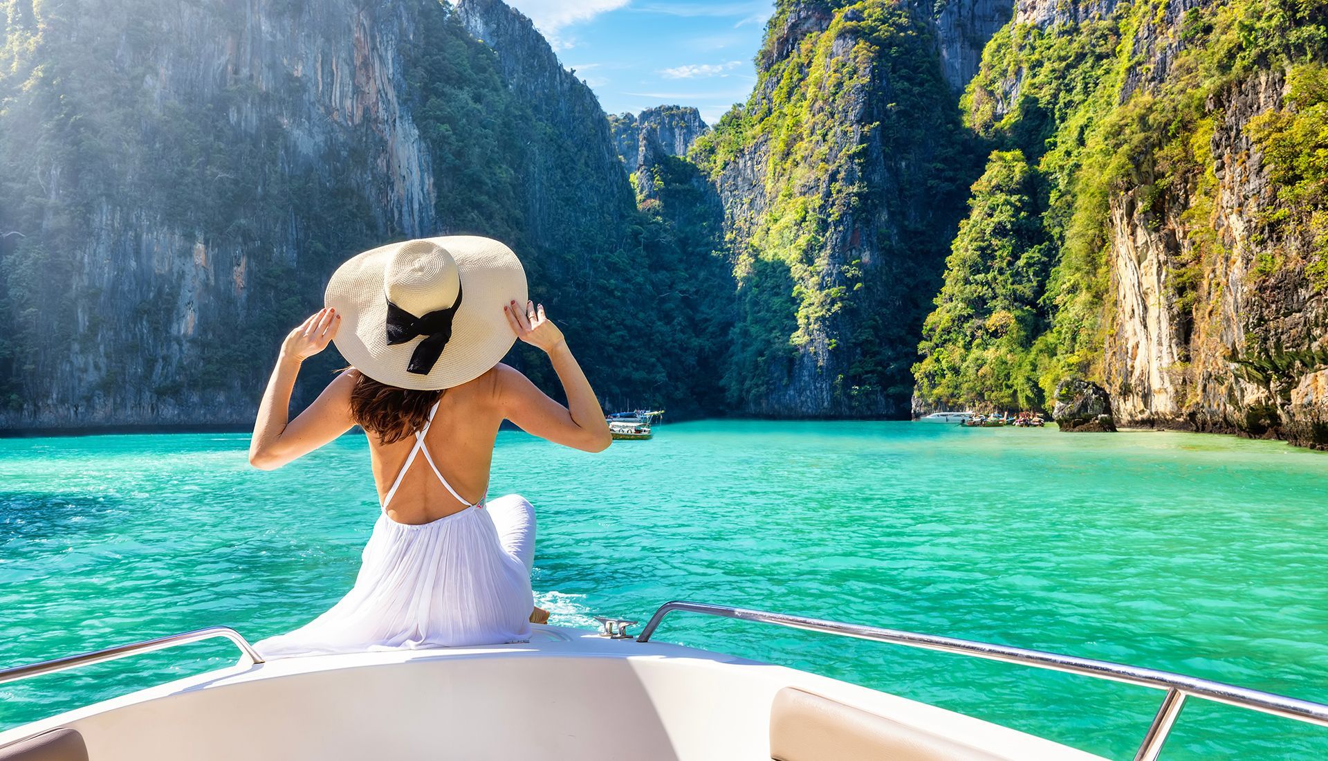 Woman on boat, looking at turquoise water and green mountains, wearing a white dress and sun hat.