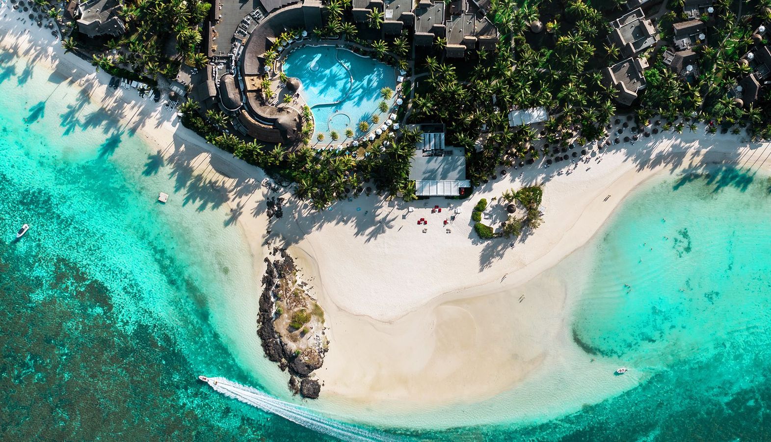 Aerial view of a resort with a turquoise pool, white sand beach, and a small boat traveling through clear blue waters.