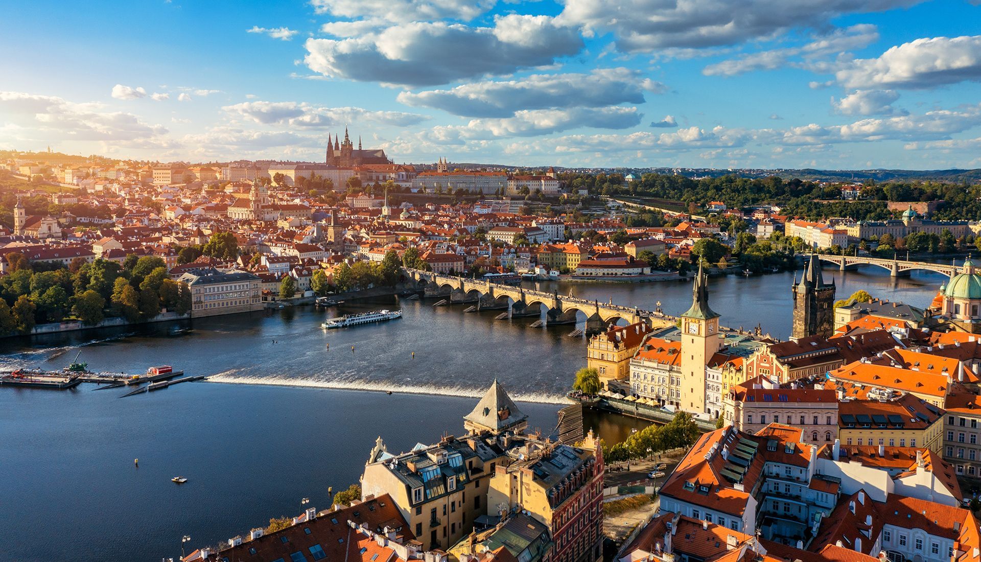 Aerial view of Prague with the Vltava River, Charles Bridge, and Prague Castle under a sunny sky.