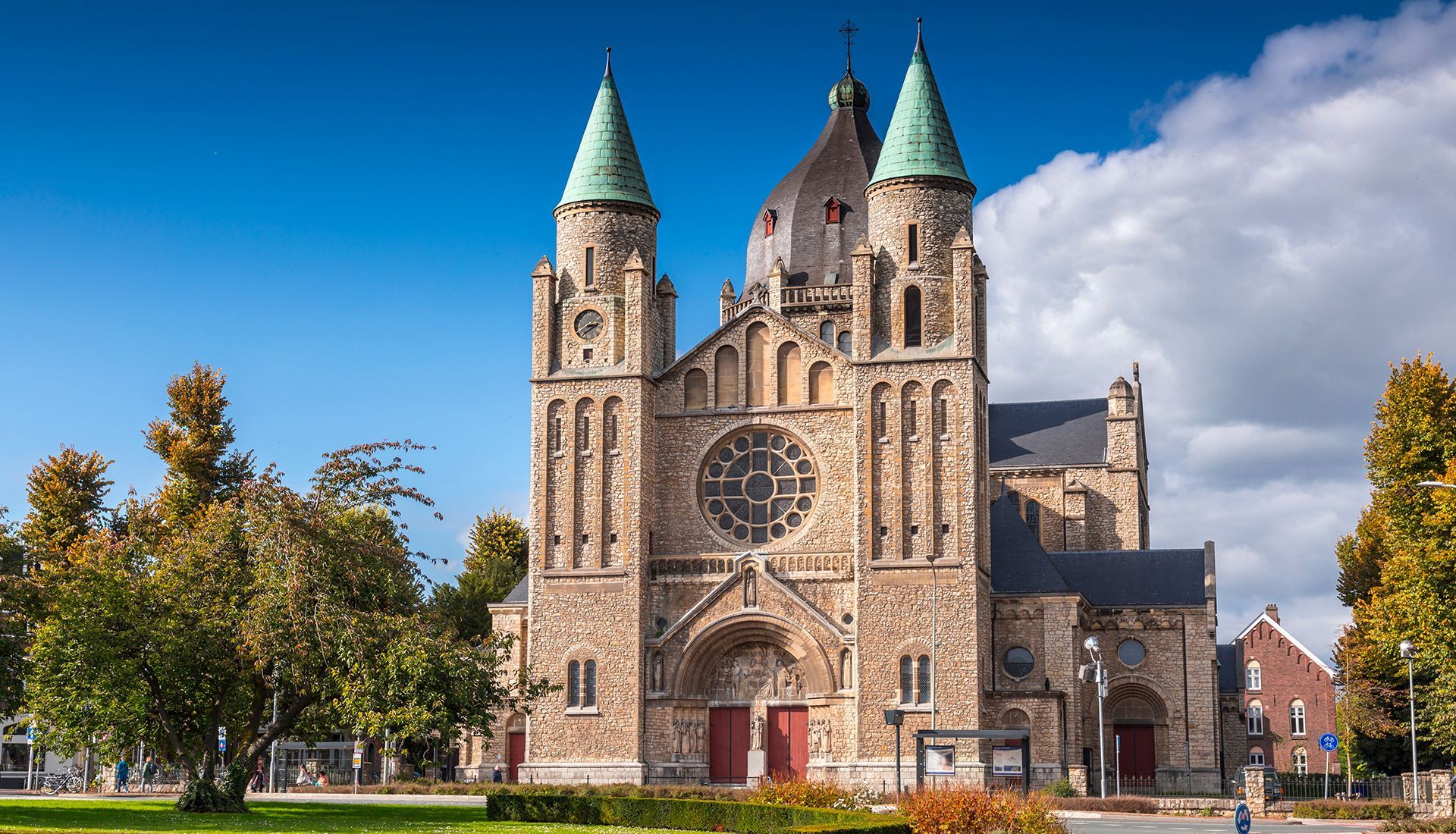 Stone church with two green-capped towers, a large rose window, and red doors against a blue sky.
