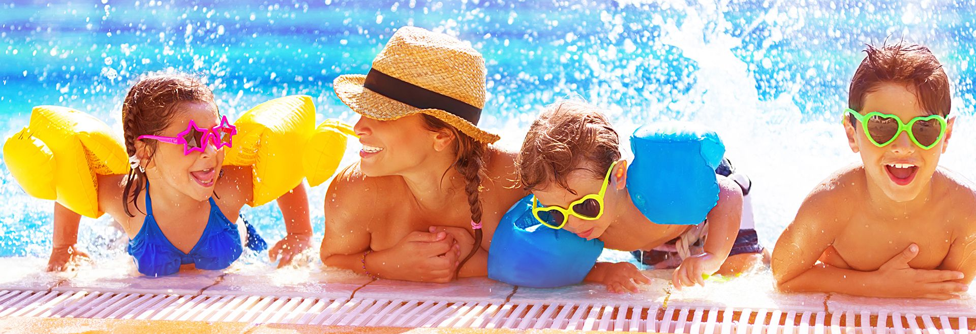 People in a pool, splashing. Woman in hat, children with arm floats and sunglasses smile. Blue water.