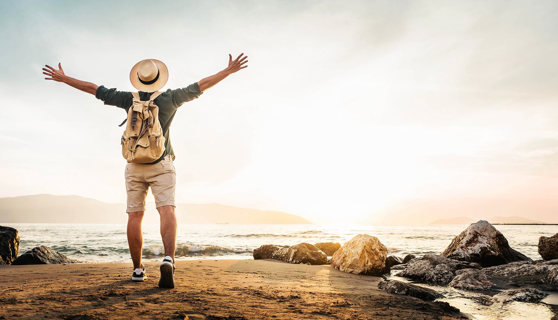 Person with arms outstretched, backpack, and hat on beach at sunset.
