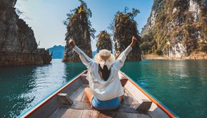Woman on boat, arms raised, on turquoise water surrounded by rock formations.