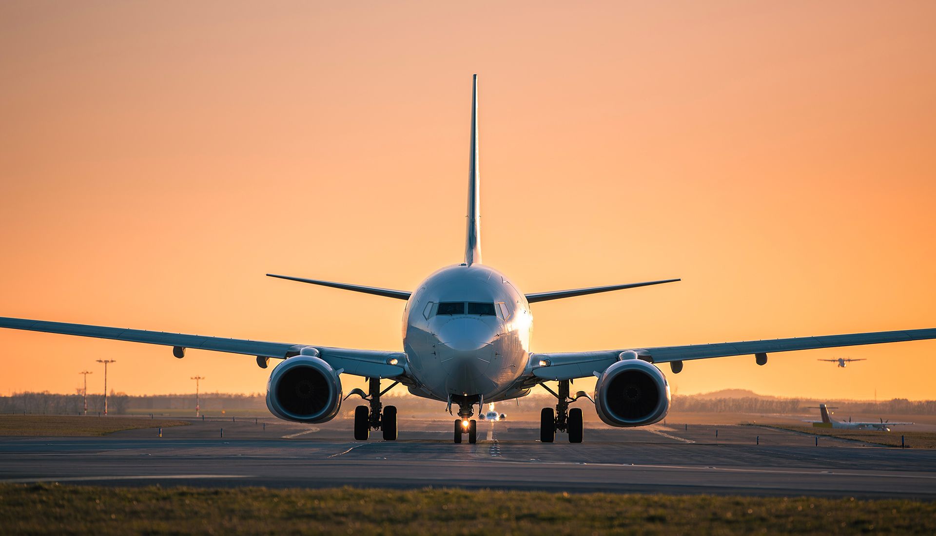 Airplane on a runway at sunset, engine and nose visible, against an orange sky.
