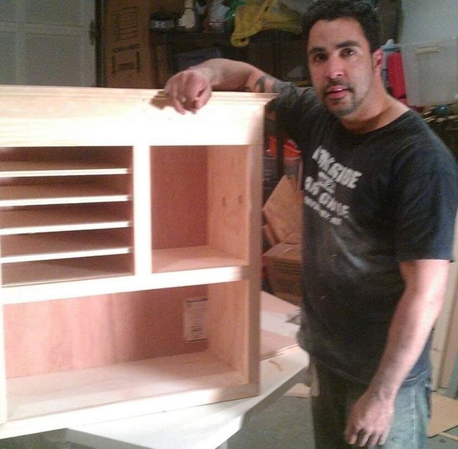 Man beside a wooden storage cabinet with shelves and slots, indoor workshop setting.