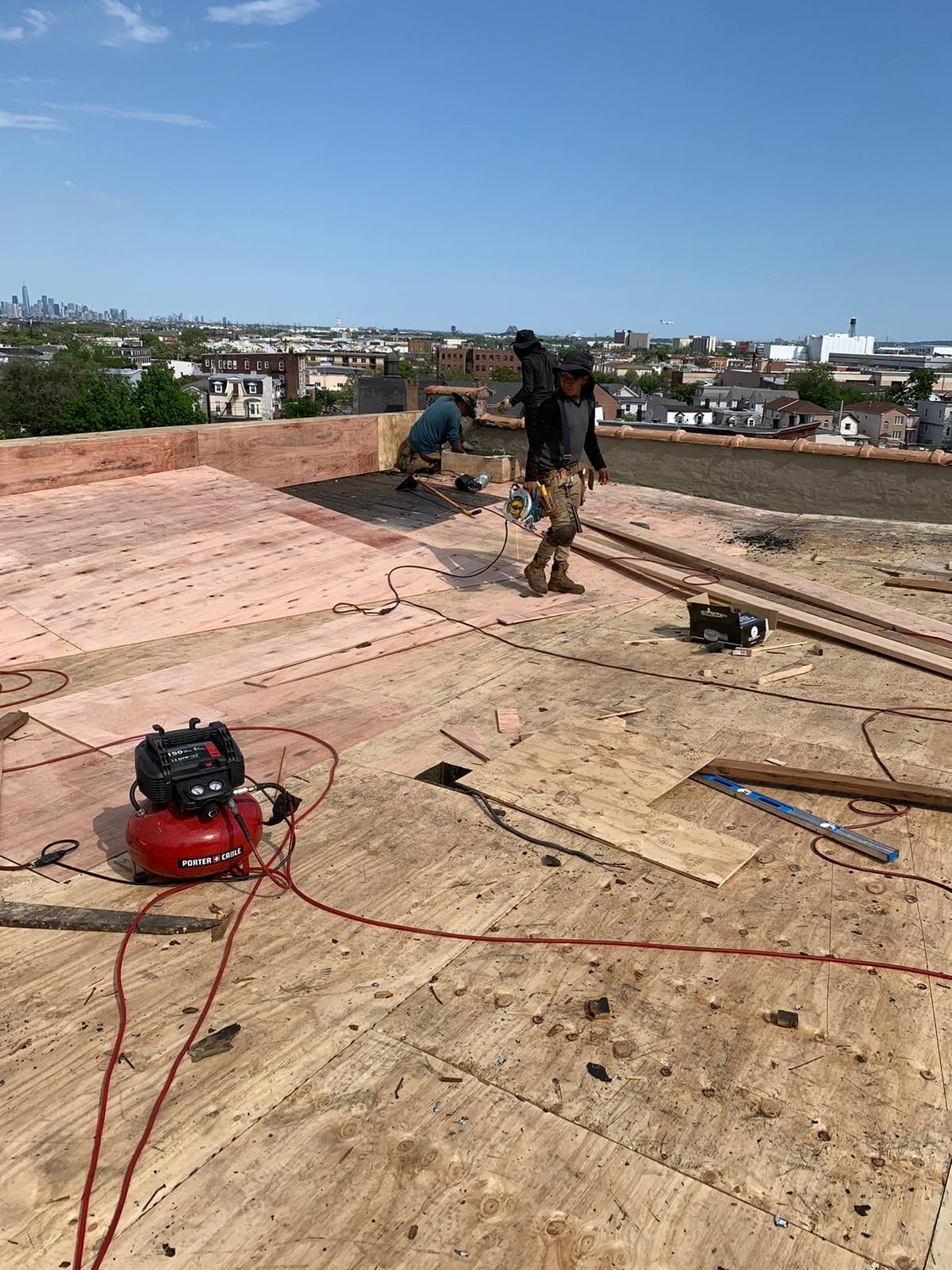 Two roofers working on a plywood roof, using tools. Red air compressor in foreground. Sunny day.
