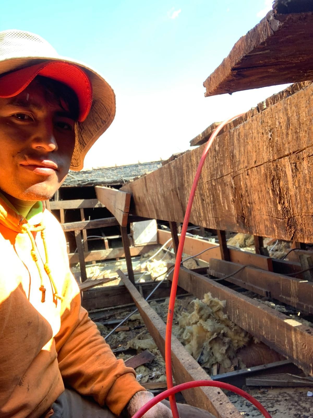 Man in orange and green work attire on a damaged roof; holding a red cord, sunlit, focused expression.