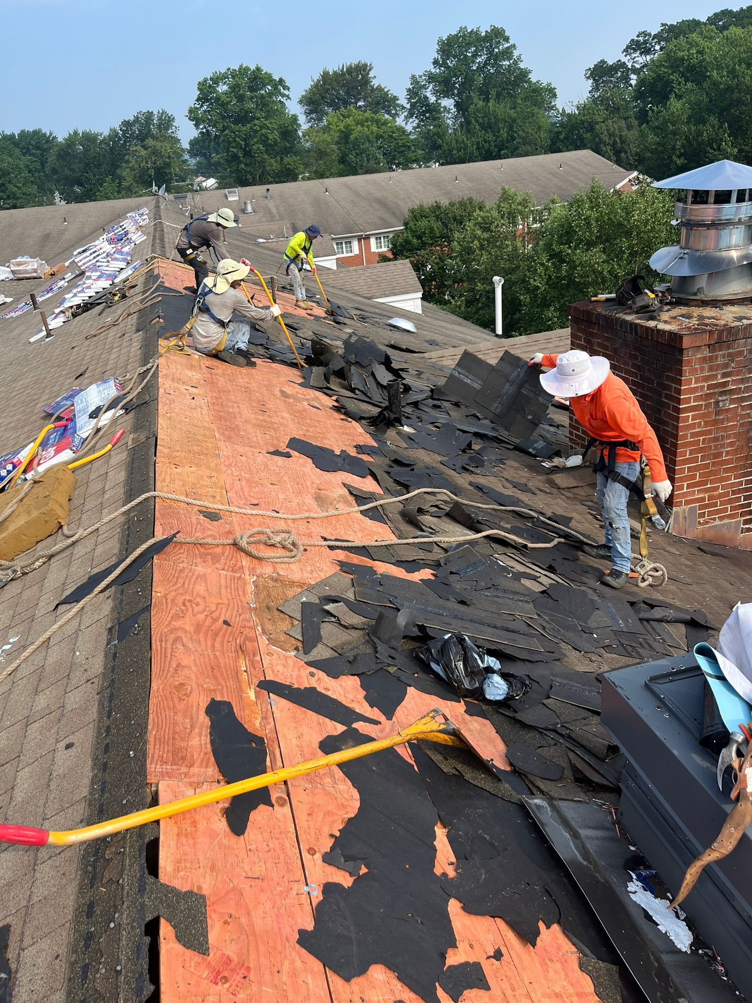 Workers removing old roofing shingles on a rooftop under a sunny sky.