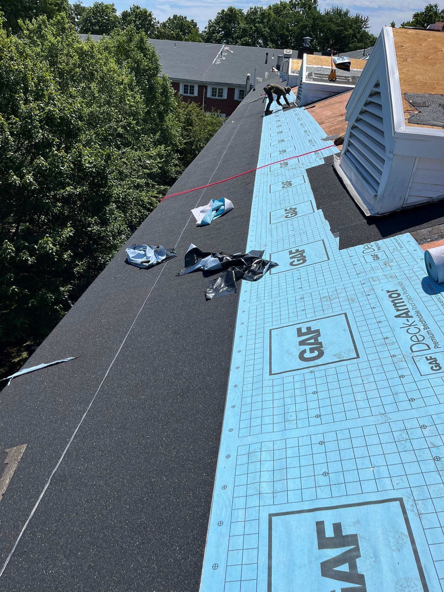 Roofers installing shingles on a residential roof, sunny day. Blue underlayment, black shingles, GAF logo visible.