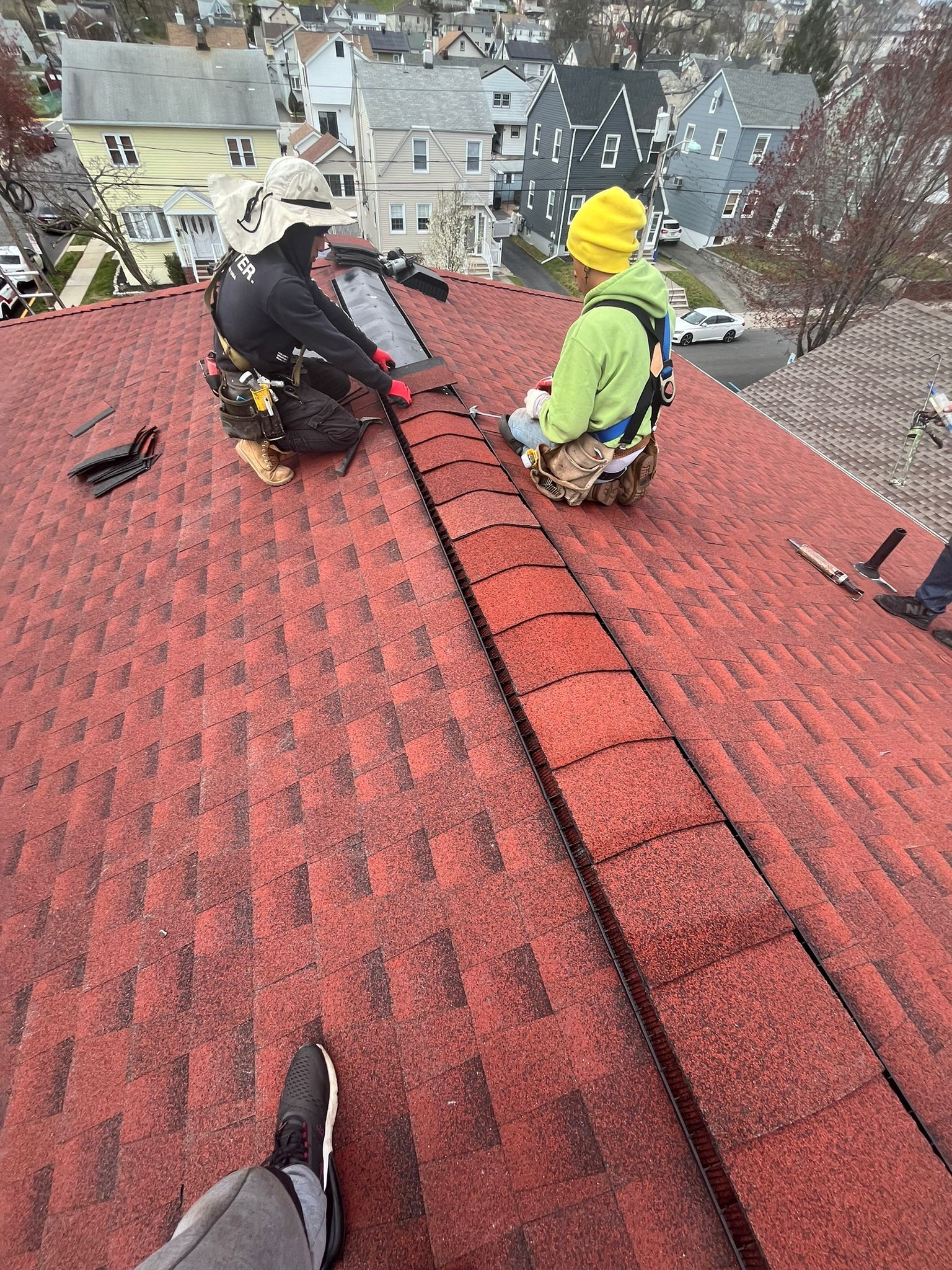 Two roofers repairing a red shingle roof, wearing safety harnesses and hard hats.