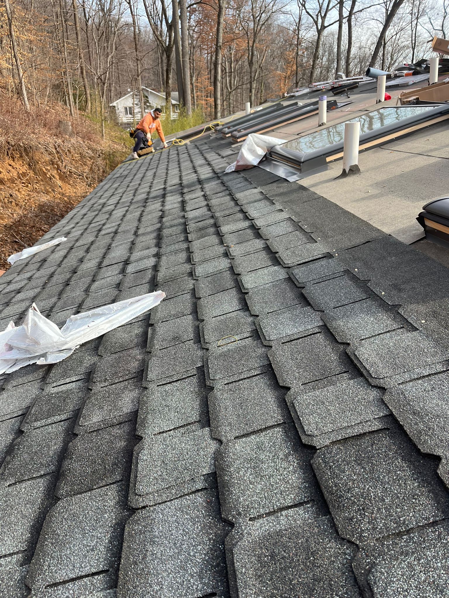 Gray shingle roof with workers in the background.  Building in a wooded area on a sunny day.