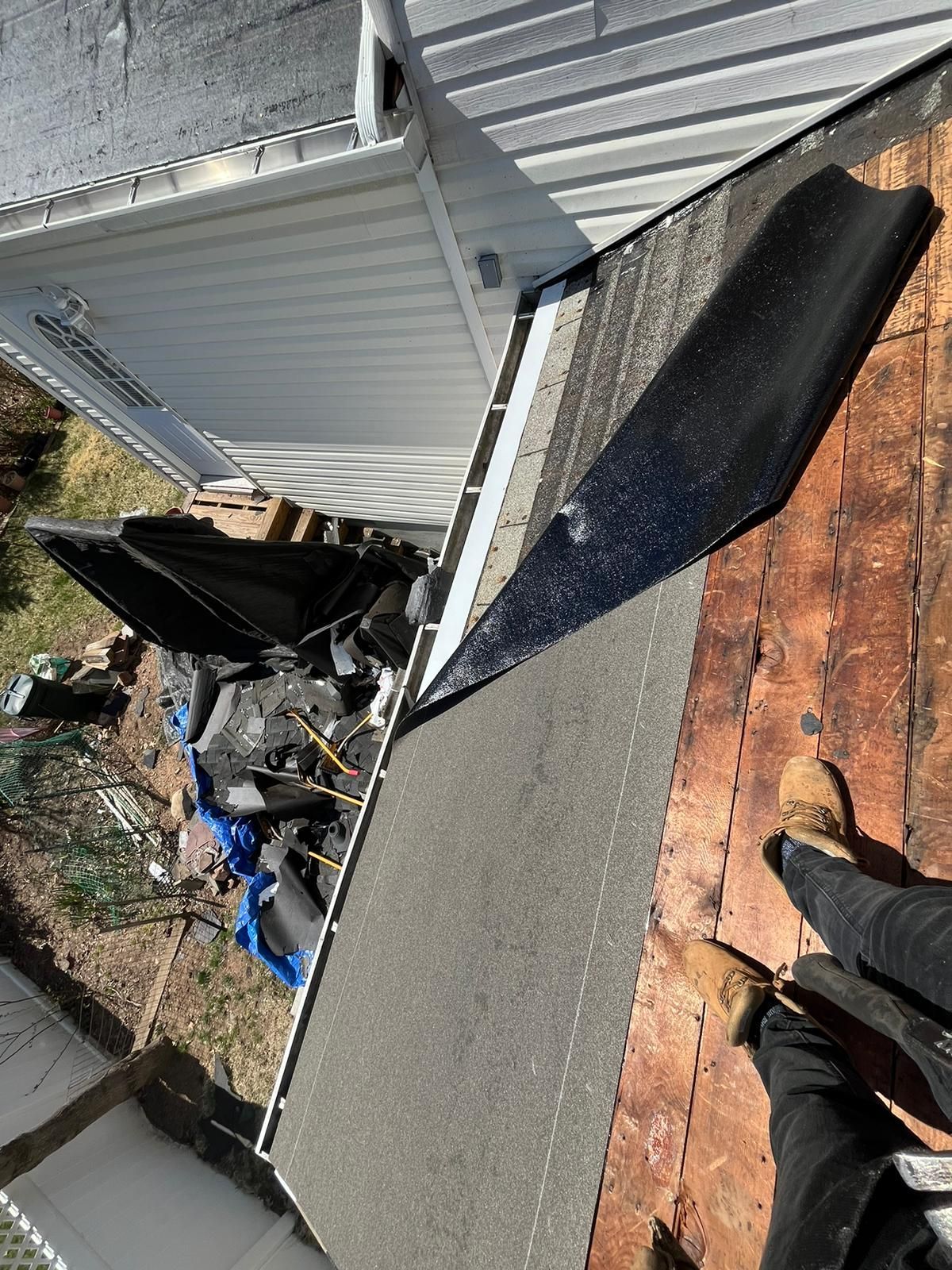 A person on a roof with a partially removed section. Debris and brown wood is visible, with a white building in the background.