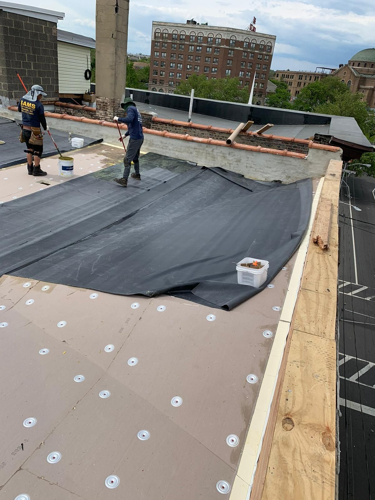 Two workers installing roofing on a building. One unrolls black membrane, other works with a tool. City backdrop.