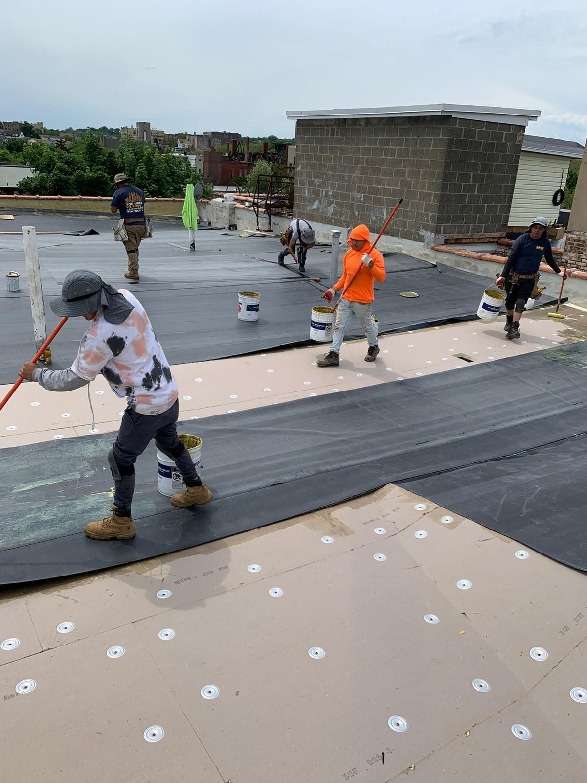 Construction workers on a roof applying sealant. Workers use rollers, wear work clothes, and work under cloudy skies.
