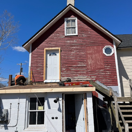 A red two-story house undergoing renovation. A person on the roof and a white door are visible.