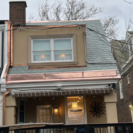 A two-story brownstone building with copper accents and a slate roof; a storefront below with a sun decoration.