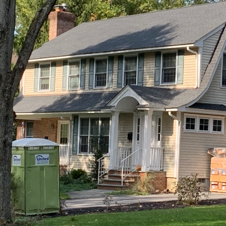 Tan two-story house with green shutters, a green portable toilet, and construction materials.