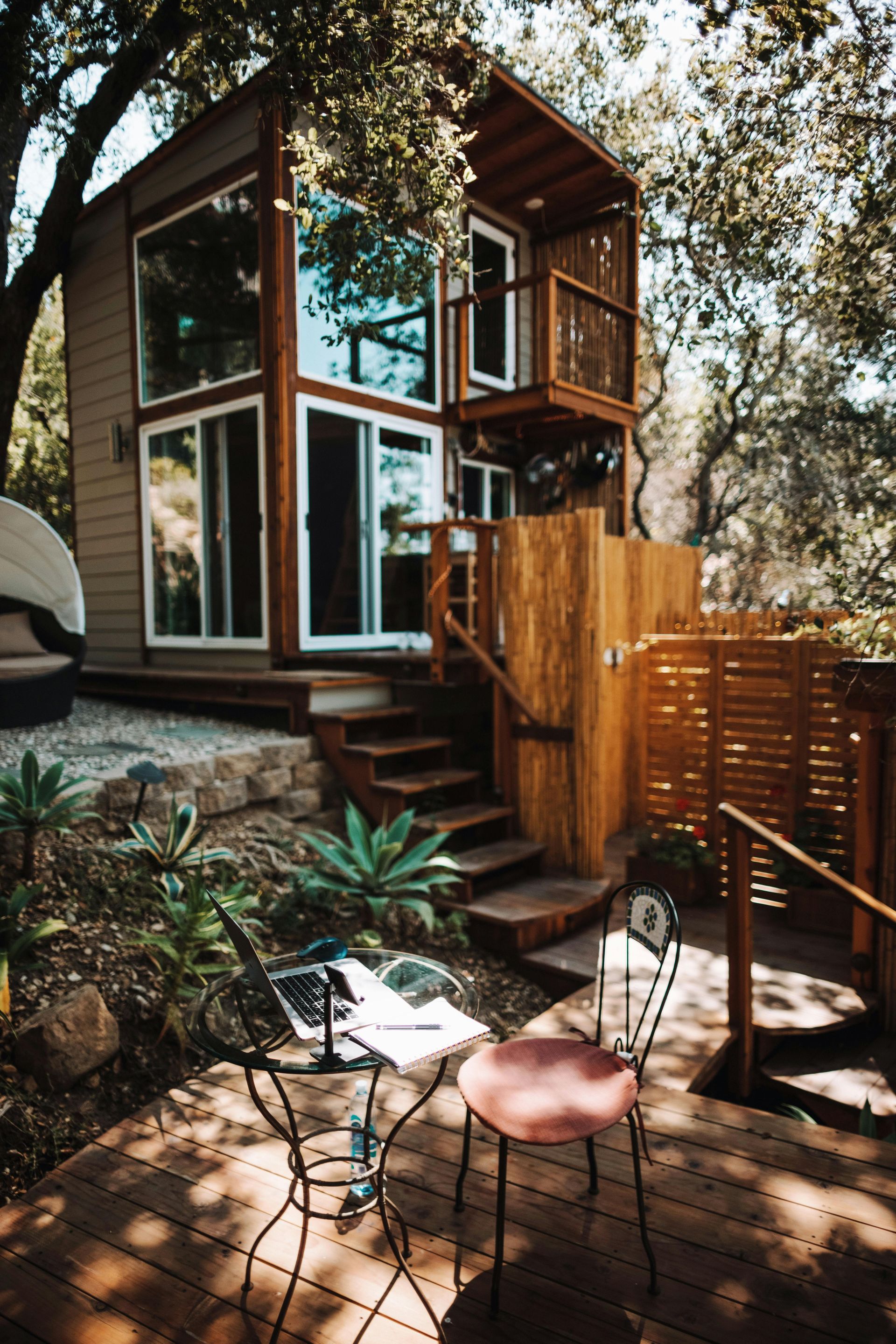 A modern two-story tiny house nestled in a wooded area, with a small outdoor patio featuring a bistro table and chair.