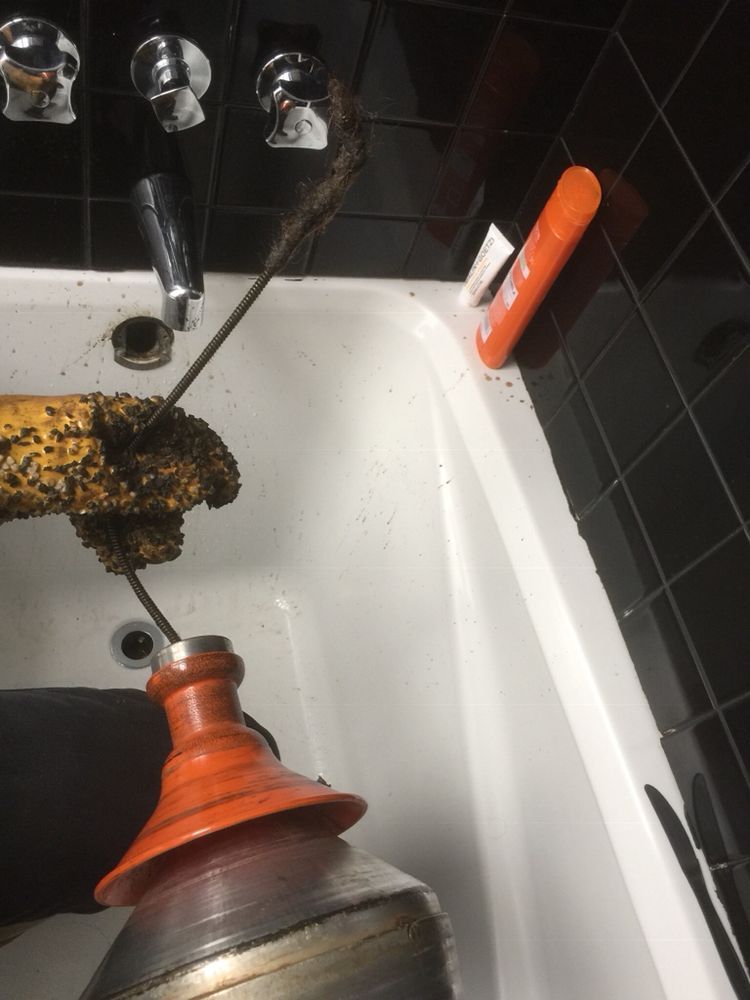 Plumber using drain snake in a white bathtub, clearing a clog. Dark hair, black tile, orange plunger visible.